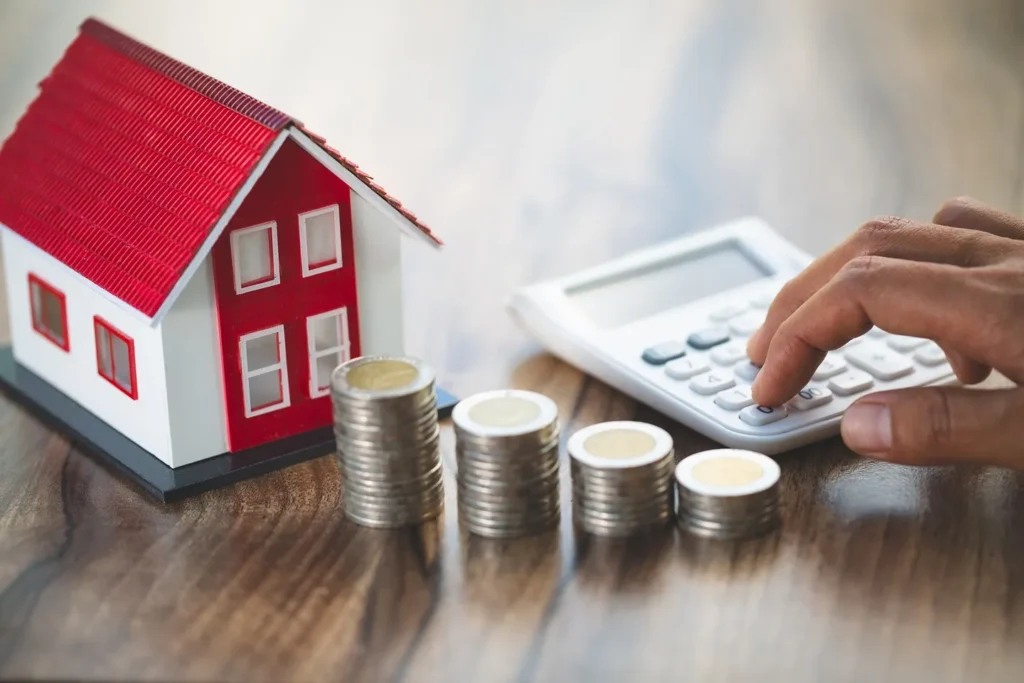 Using a calculator next to stacks of coins and a toy house.