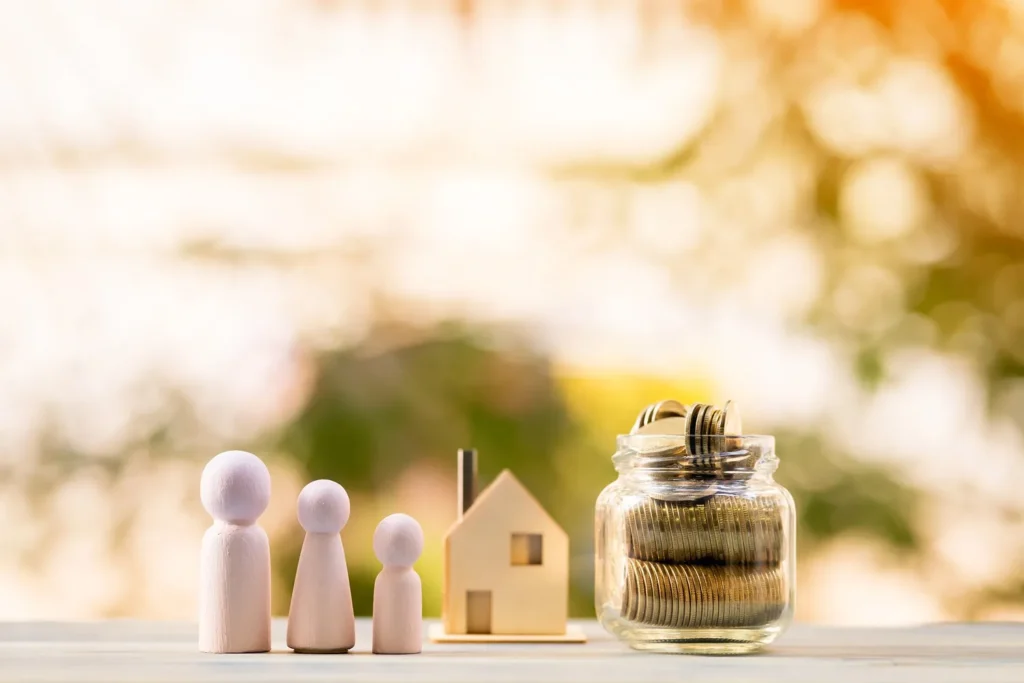 Wooden family figurines, a wooden model home and a jar of coins on a table.