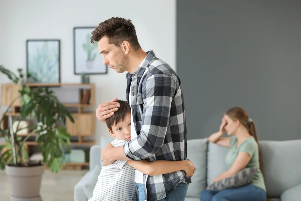 A young child hugs his dad while a mom sits on the couch holding her head.